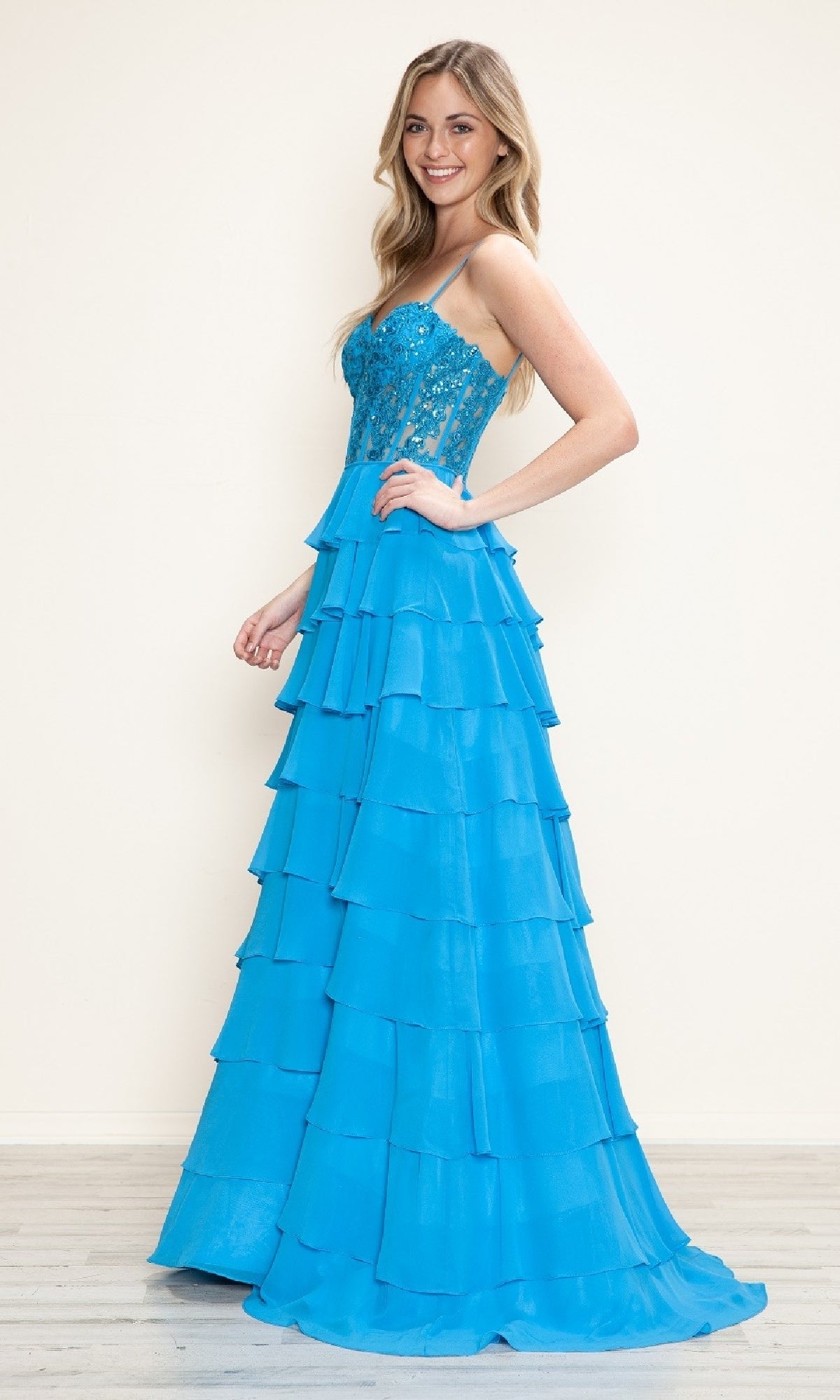 A woman stands in profile, smiling, in the Long Tiered Corset Prom Dress: Poly USA 9698—a bright blue a-line prom dress with spaghetti straps and a corset bodice—set against a plain light background.